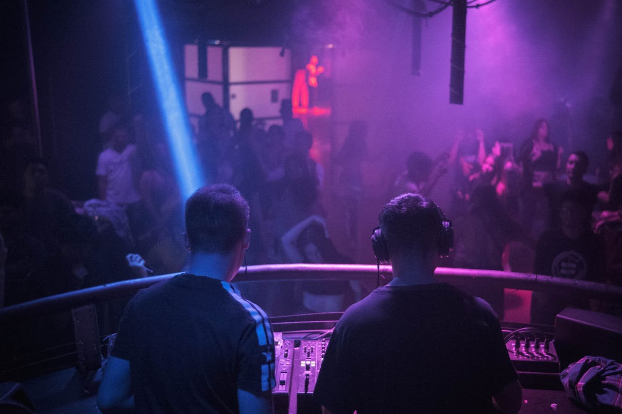 our-story A lively night club scene with a DJ mixing music amidst vibrant lights and dancing crowd.